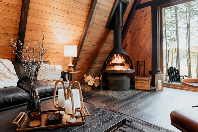 Intérieur d'un chalet en bois au style scandinave, avec un poêle à bois noir allumé, dégageant une ambiance chaleureuse. Un canapé noir avec des coussins blancs se trouve à gauche, une table basse en bois foncé est ornée de bougies et d'un vase avec des branches. Une grande fenêtre offre une vue sur une forêt, laissant entrer la lumière naturelle.